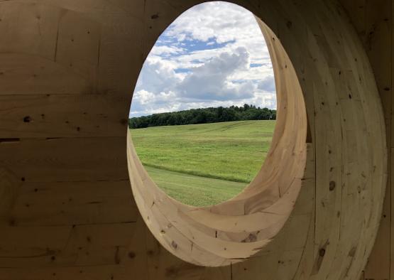 a hole in a wood wall with a field of grass and clouds